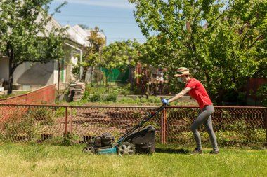 Bahçesinde çim biçme makinesiyle çim biçen bir kadın güneşli bir günde Coronavirus salgını yüzünden cerrahi maske takıyor..