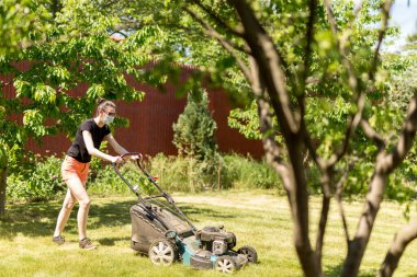 Bahçesinde çim biçme makinesiyle çim biçen bir kadın güneşli bir günde Coronavirus salgını yüzünden cerrahi maske takıyor..