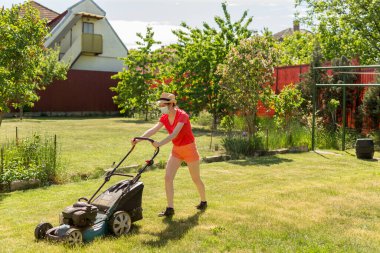 Bahçesinde çim biçme makinesiyle çim biçen bir kadın güneşli bir günde Coronavirus salgını yüzünden cerrahi maske takıyor..