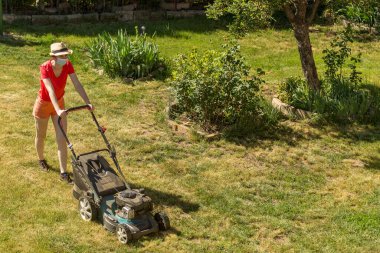 Bahçesinde çim biçme makinesiyle çim biçen bir kadın güneşli bir günde Coronavirus salgını yüzünden cerrahi maske takıyor..