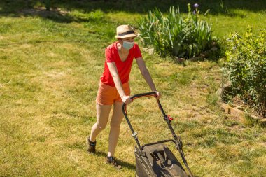 Bahçesinde çim biçme makinesiyle çim biçen bir kadın güneşli bir günde Coronavirus salgını yüzünden cerrahi maske takıyor..