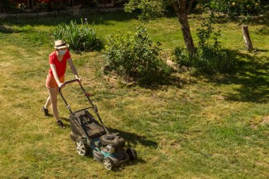 Bahçesinde çim biçme makinesiyle çim biçen bir kadın güneşli bir günde Coronavirus salgını yüzünden cerrahi maske takıyor..