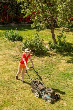 Bahçesinde çim biçme makinesiyle çim biçen bir kadın güneşli bir günde Coronavirus salgını yüzünden cerrahi maske takıyor..