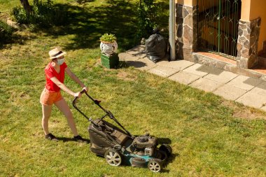 Bahçesinde çim biçme makinesiyle çim biçen bir kadın güneşli bir günde Coronavirus salgını yüzünden cerrahi maske takıyor..