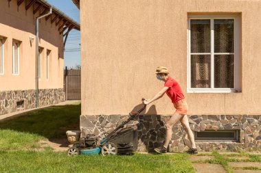 Bahçesinde çim biçme makinesiyle çim biçen bir kadın güneşli bir günde Coronavirus salgını yüzünden cerrahi maske takıyor..