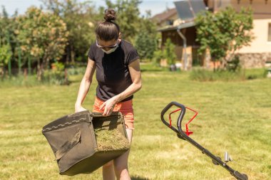 Bahçesinde çim biçme makinesiyle çim biçen bir kadın güneşli bir günde Coronavirus salgını yüzünden cerrahi maske takıyor..