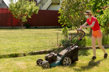Bahçesinde çim biçme makinesiyle çim biçen bir kadın güneşli bir günde Coronavirus salgını yüzünden cerrahi maske takıyor..