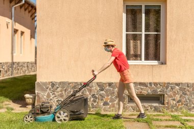 Bahçesinde çim biçme makinesiyle çim biçen bir kadın güneşli bir günde Coronavirus salgını yüzünden cerrahi maske takıyor..