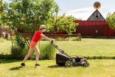 Bahçesinde çim biçme makinesiyle çim biçen bir kadın güneşli bir günde Coronavirus salgını yüzünden cerrahi maske takıyor..