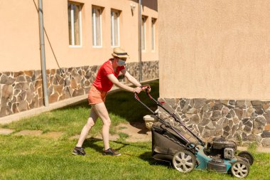 Bahçesinde çim biçme makinesiyle çim biçen bir kadın güneşli bir günde Coronavirus salgını yüzünden cerrahi maske takıyor..