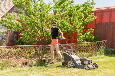 Bahçesinde çim biçme makinesiyle çim biçen bir kadın güneşli bir günde Coronavirus salgını yüzünden cerrahi maske takıyor..