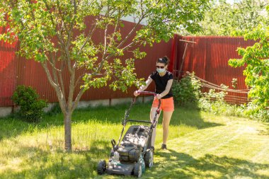Bahçesinde çim biçme makinesiyle çim biçen bir kadın güneşli bir günde Coronavirus salgını yüzünden cerrahi maske takıyor..