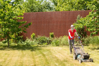Bahçesinde çim biçme makinesiyle çim biçen bir kadın güneşli bir günde Coronavirus salgını yüzünden cerrahi maske takıyor..