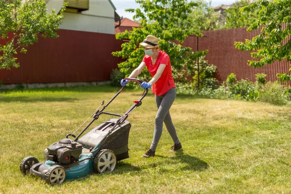 家で晴れた日に芝生の芝刈り機で草を刈る彼女の裏庭の女性は コロナウイルスの流行のために外科用マスクを着て ストック画像