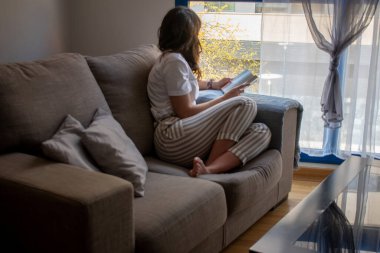 Genç bir kadın kanepesinde oturmuş kitap okuyor ve çay içiyor.