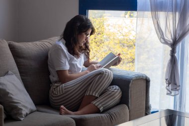 Genç bir kadın kanepesinde oturmuş kitap okuyor ve çay içiyor.