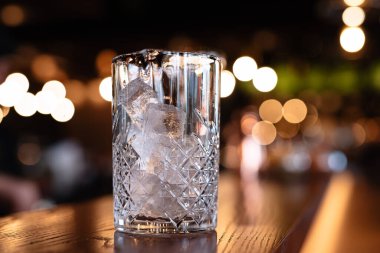 Kristal şeffaf cam buzla dolu, bardaktaki buz küplerin içinde, bir bar tezgahında, kahverengi bir masa, arka plan bulanık ve aydınlık ve parlak.