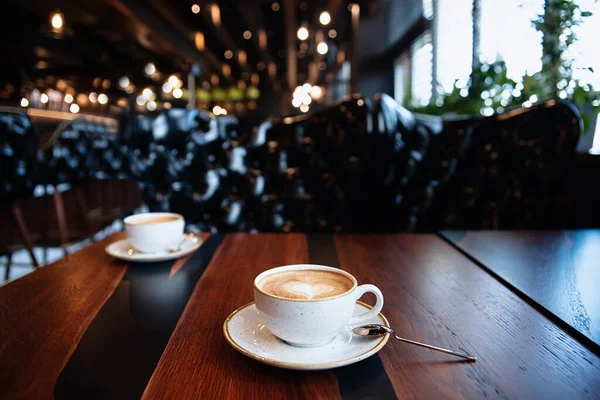 Masanın üzerinde iki fincan kahve, restoran, kahverengi mobilya, loş ışıklar, bir beyaz kupa ve bir tabakta metal kaşık, yakın plan, kapuçino, kahverengi ahşaptan yapılmış bir masa, seramik, kil tabaklardan yapılmış., 