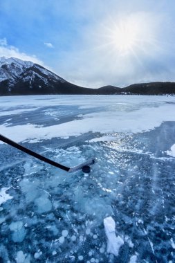 Kanada, Alberta 'daki Banff Ulusal Parkı' nın Kanada kayalıklarında Minnewanka Gölü 'nde buz hokeyi oynuyorum. Buz yüzeyinin altında donmuş metan gazı kabarcıkları olan hokey sopası ve disk..