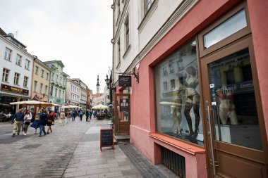 İnsanlar eski şehrin sokakları boyunca yürüyorlar. İç çamaşırlı manikinlerin olduğu bir vitrin. Eski kule uzaktan görülebilir. güzel renkli evler