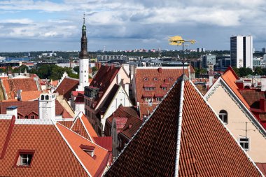 Bulutlu mavi gökyüzüne karşı horoz şeklinde bir altın rüzgar gülü ve altında eski bir Avrupa şehrinin kırmızı ve turuncu üçgen kiremitli çatıları vardır. Uzakta eski kulenin kulesi
