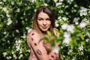 Güneşli bir bahar gününde pembe elbiseli güzel bir kızın portresi. Çiçek açan elma bahçesindeki beyaz çiçeklerin arka planında bize bakıyor..