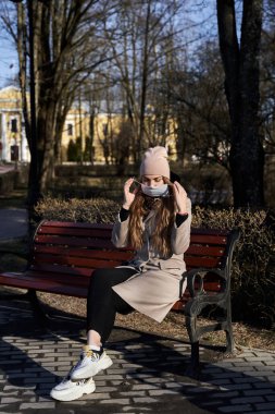 Şapkalı ve ceketli sarışın, tıbbi koruyucu bir maske takıyor. Kız beyaz ayakkabılarıyla parkta bir bankta oturuyor.