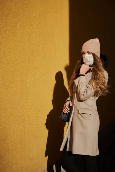 医療保護マスクの美しいブロンドの女の子は太陽の光の下で通りの黄色い壁に面して立っている彼女はあごの下に手を握っている コートとかバッグとか帽子とか — ストック写真