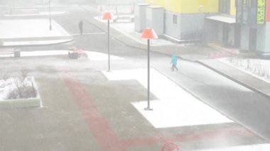 Mavi, kalın kıyafetler giymiş bir çocuk kar fırtınası sırasında bir futbol topunu tekmelerken karlı bir yolda koşuyor. Çevrelerinde, renkli evler var.