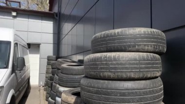 Birçok eski kullanılmış lastik gri bir duvarın yanında duruyor. Yanında geri dönüşüm ve geri dönüşüm için onu alacak büyük beyaz bir araba var.