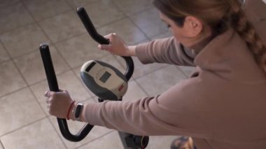 Ceketli bir kız spor spor bisikletinin ve pedalların boynuzlarını tutuyor. Parametreler ekranda gösterilir. Elinde bir spor bilekliği var. Örgülü sarı saçlar..