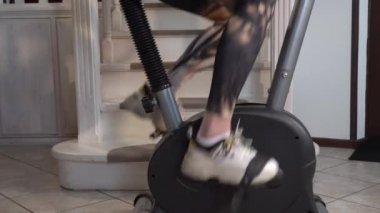 Beyaz spor ayakkabılarla güzel kadın bacaklarının yakın çekimi ve dar spor taytları ev egzersiz bisikletinin pedallarını çeviriyor. Metal boruları olan gri bir simülatör merdivenlerin yanında duruyor.
