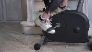 Beyaz spor ayakkabılı güzel kadın bacakları ve dar spor taytları ev egzersiz bisikletinin pedallarını büküyor. Metal boruları olan gri bir simülatör merdivenlerin yanında duruyor.