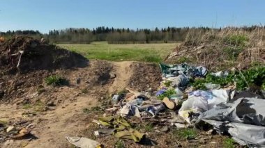 Eski çöp yığınları, paçavralar, ayakkabılar, plastik, film, lastik, tek kullanımlık eşyalar doğaya atılır. Yeşillik bir çayırın yanında, genç yapraklı ağaçlar, tarlalar ve mavi gökyüzü. Yasadışı çöplük