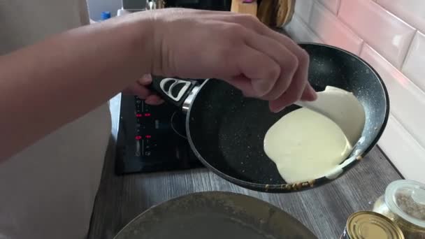 les mains féminines verser une cuillerée de pâte dans une poêle chaude pour faire frire les crêpes. met une poêle sur une cuisinière électrique. au loin est une pile de crêpes prêtes à l'emploi dans la cuisine.