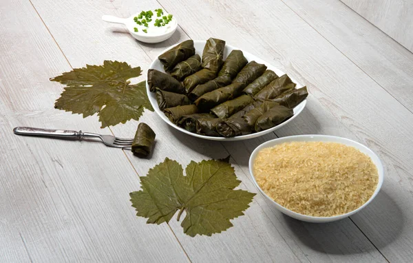 Dolma. Biftek ve pirinçle doldurulmuş üzüm yaprakları. Baharatlı beyaz sos. Bir tabak pirinç. Yan görünüş, açık ahşap bir zemine yakın.