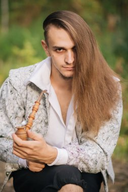 Ormanda elinde bir boru ile seksi uzun saçlı adam. Güzel, eski ceketle içinde desen ve beyaz bir gömlek. Fantezi.