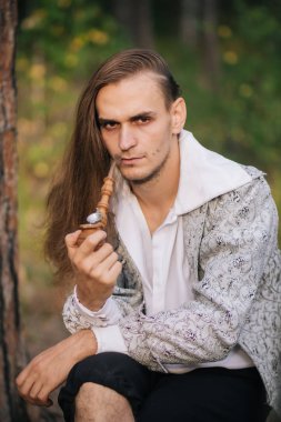 Ormanda elinde bir boru ile seksi uzun saçlı adam. Güzel, eski ceketle içinde desen ve beyaz bir gömlek. Fantezi.