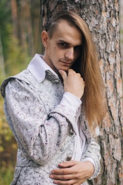 Sexy long-haired man with a pipe in his hands, in the woods. In a beautiful, ancient jacket with patterns and a white shirt. Fantasy. clipart