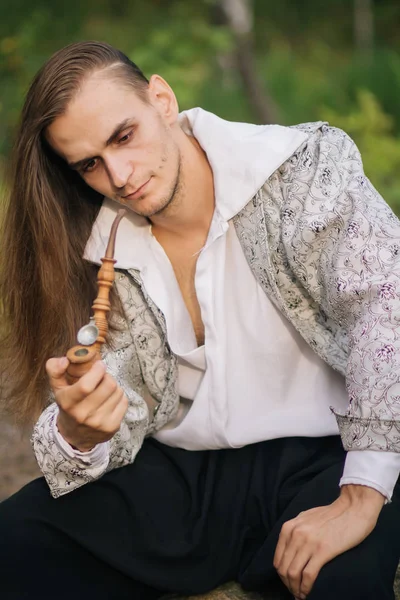 Sexy long-haired man with a pipe in his hands, in the woods. In a beautiful, ancient jacket with patterns and a white shirt. Fantasy.