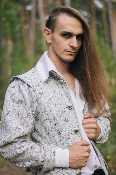 Sexy long-haired man with a pipe in his hands, in the woods. In a beautiful, ancient jacket with patterns and a white shirt. Fantasy.