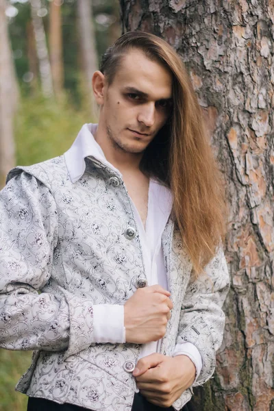 Sexy long-haired man with a pipe in his hands, in the woods. In a beautiful, ancient jacket with patterns and a white shirt. Fantasy.