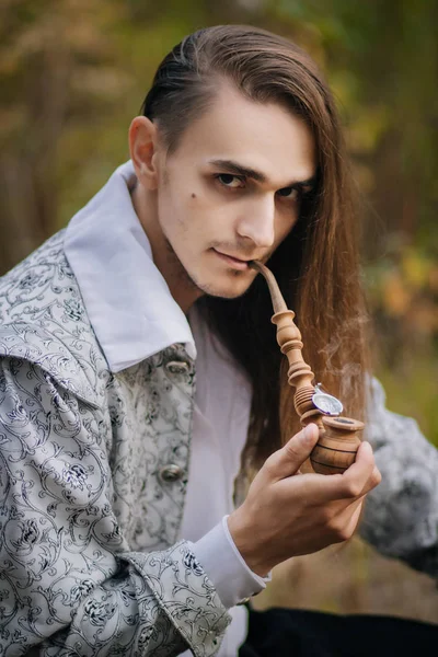 性感长发男子与烟斗在他的手 在树林里 穿着一件漂亮的古老的夹克 上面有花纹和白色衬衣 — 图库照片