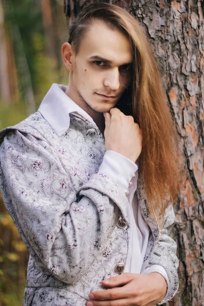 Sexy long-haired man with a pipe in his hands, in the woods. In a beautiful, ancient jacket with patterns and a white shirt. Fantasy.