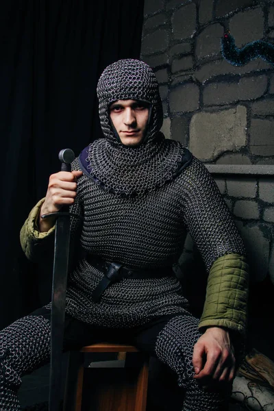 A man with a gun, a stern look. Model man in chain mail hood and chain mail with a sword in his hand. Knight in armor in the castle. Medieval photo illustrations.