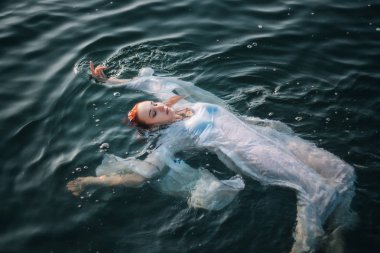 Güzel kızıl saçlı kız suda yüzüyor. Kayalık sahil. Bir model beyaz bir antika elbise zümrüt suda. Ortaçağ görüntüsü. Fantezi