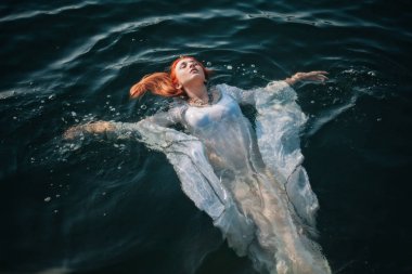 Güzel kızıl saçlı kız suda yüzüyor. Kayalık sahil. Bir model beyaz bir antika elbise zümrüt suda. Ortaçağ görüntüsü. Fantezi