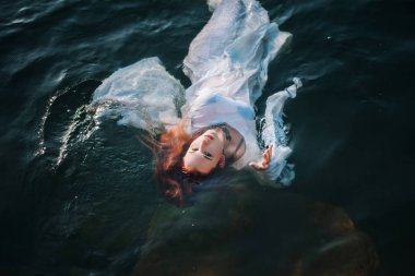 Güzel kızıl saçlı kız suda yüzüyor. Kayalık sahil. Bir model beyaz bir antika elbise zümrüt suda. Ortaçağ görüntüsü. Fantezi