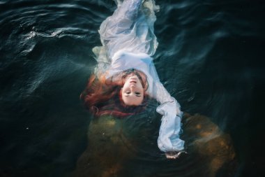 Güzel kızıl saçlı kız suda yüzüyor. Kayalık sahil. Bir model beyaz bir antika elbise zümrüt suda. Ortaçağ görüntüsü. Fantezi