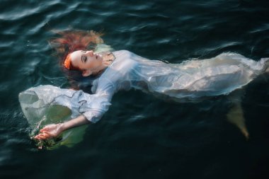 Güzel kızıl saçlı kız suda yüzüyor. Kayalık sahil. Bir model beyaz bir antika elbise zümrüt suda. Ortaçağ görüntüsü. Fantezi